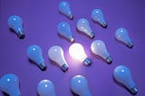 This image shows a collection of light bulbs on a purple background. All bulbs are turned off except for one in the center, which is illuminated, creating a focal point among the unlit bulbs surrounding it.