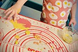 Photo shows the torso of a child wearing a pink shirt with images of fried eggs on it. On the table in front of her is a mat with blobs of pink and yellow goop on it, and she’s dipping a finger into the yellow.