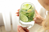 Woman’s hands holding a mason jar full of water with a whole cucumber sliced in