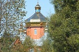 A brick church tower with a shining metal Orthodox dome, surrounded by trees.
