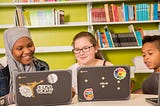 Three students looking at laptops and smiling in front of a bookcase.
