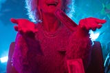 Image description: An old woman with sunglasses wearing a lacy dress is lit in red light with money raining down on her