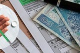 Na zdjęciu znajdują się dokumenty podatkowe PIT, polskie banknoty o nominałach 100 i 50 złotych oraz czarny długopis. Na pierwszy plan wysuwa się również okrągłe wstawienie z dłonią trzymającą różnokolorowe kapsułki i tabletki. Kompozycja obrazu podkreśla finansowe aspekty leczenia, sugerując konieczność rozliczania wydatków na leki i zwrot za leki dla niepełnosprawnych. Zdjęcie oddaje poczucie formalności, biurokracji oraz konieczności uważnego planowania budżetu na zdrowie.