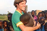 Smiling white man playfully holding a black African child as other black children look on, smiling, in the background.