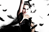 An image shows Natalie Portman in a black gown. She has black eyeshadow and a sparkling white tiara on her head. She stands with one hand in the air and one on her waist. Black feathers fly all around her.