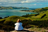 A woman in a white jacket and hat faces away from the camera and overlooks the Otago Bay and Peninsula in New Zealand, which has lush blue water and a green landscape.