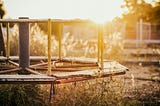 A rusty, old-fashioned merry-go-round rots at a derelict, overgrown playground against a sunset background.