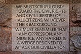 Stone memorial plaque with engraved text reading: ‘WE MUST SCRUPULOUSLY GUARD THE CIVIL RIGHTS AND CIVIL LIBERTIES OF ALL CITIZENS, WHATEVER THEIR BACKGROUND. WE MUST REMEMBER THAT ANY OPPRESSION, ANY INJUSTICE, ANY HATRED, IS A WEDGE DESIGNED TO ATTACK OUR CIVILIZATION.’ The text is carved into reddish-brown granite or sandstone blocks with visible mortar lines between them.
