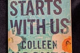 A paperback copy of “It Starts With Us” by Colleen Hoover. The cover is a pastel blue-aqua color showing flowers below the title and highlighting the author name. At the top center, there is a yellow sticker that says “Every ending has a beginning.” At the top right, there is a Barnes and Noble “Buy 1, Get 1 50% Off” sticker. Under the author name, the text says “#1 New York Times Bestselling Author of “It Ends With Us.”