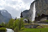 a quaint pretty view of a little hamlet nestled in the mountains, with a waterfall and a brook flowing next to it