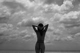 A grayscale photograph of a skinny woman looking away from the camera. She has straight black hair and is putting her hands behind her head. She is staring out into the ocean and many white clouds appear in the sky. (2024)