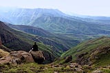 Women seen from behind sitting with crossed legs on rock while overlooking lush, green mountain valley