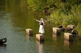 Dressed in white, woman stands on concrete pylons in Bronx River and gestures across the water, one finger pointing forward