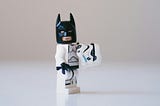 Close up of a Lego storm trooper wearing a batman mask and holding its helmet on a blurry white background.