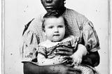 Black and white photograph of black woman wearing a long checkerd dress holding a white child from 1858.