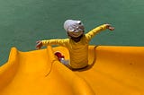 Little boy wearing a yellow shirt and gray hat sliding down a yellow slide with open arms.