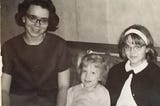 Black and white photo of three white girls: two with cat’s eye glasses. One has dark hair and is about 18; one is blonde haired and about 5 or 6; and the third is about 12 or 13, has dark blonde hair and wears a white headband. All are smiling.