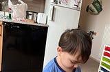 One-year-old Gabriel’s attention is captured entirely by the task of peeling a clementine. He wears a blue long-sleeved shirt adorned with a green dinosaur, and his hair is playfully tousled.