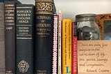 A photo of some English grammar and and usage guides, including some hardbacks (e.g., “Fowler’s Modern English Usage” and a dictionary of classical quotations) and softbacks (e.g., Strunk and White’s “The Elements of Style”). A quote in handwritten calligraphy on a small card sits to the right: “There are only four subjects in the curriculum of life — love, justice, courage, and compassion. — Richard Trelease.” (Photo by author.)