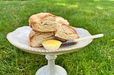 A cream colored gold-rimmed cake stand with four cream scones on it, a small pearl-handled knife, and a carved stone white bowl full of mango curd out on the grass of the garden.