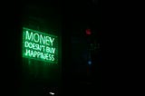 The image shows a green neon sign displayed in a dark room that reads “MONEY DOESN’T BUY HAPPINESS” in bright, glowing letters. The sign is mounted on what appears to be a wall, creating a striking contrast against the black background. To the right of the frame, there’s a red EXIT sign visible in the darkness. The atmosphere is moody and contemplative, with the neon green light casting a subtle glow on the surrounding area.