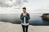 Man in blue denim jacket standing standing on cliff near body of water