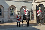 Three soldiers dressed in ceremonial uniforms, one standing on the ground and two sitting on horses.