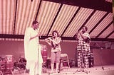 Three people on a stage playing the flute. There is a white woman in middle and two African men in traditional African dress to either side of her.