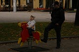 Man in his 30s or 40s with hands in pockets uses his foot to rock a playground horse on a spring with his child of 3 or 4 in the rider’s seat.