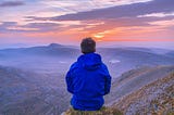 A guy sitting on the cliff of a mountain watching the clouds and a most beautiful sunset.