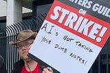 A picketer carries a sign reading “Writers Guild on Strike! A.I.’s not taking your dumb notes!”