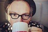 Selfie of author wearing vintage spectacles, bowler and a black-and-white polka dot shirt sips coffee from a TED mug