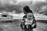 Baby looking at the camera while woman holding it looks behind her toward a trailer.