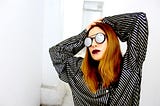 A woman in round-framed sunglasses and a striped shirt strikes a pose with her hands on her head.