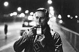 A man stands on the side of a city street at night wearing a leather jacket and taking a breathalyzer test