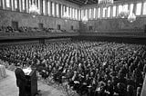 A large ballroom filled with people seated in rows, listening to a man at lectern.