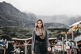 Aelegant lady walks courageously forward. Behind her is a hilly and foggy background. The hills have a thick forest covering and below them some wooden structures in what appears to be a small town.