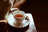 Pouring orange-colored tea into a small cup from a classic kettle. Next to the tea cup is a silver spoon of normal size.