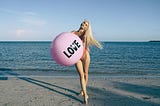 woman in swimsuit on the beach holding a large pink ball that says “love” on it