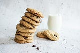 A tower of chocolate chip cookies leans towards a glass of milk and couple more delicious cookies.