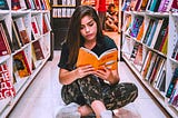 color photpgraph of a young woman reading a book with an orange cover with sitting cross-legged on the floor between two full shelves of books in the library