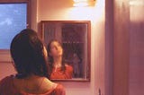 A young woman wearing an orange shirt stands in front of a bathroom mirror, her hair wet from a shower. The reflection is blurry and she is gazing upwards past the top right corner of the frame, conveying that she is contemplating something.