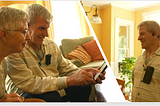 An older man and woman look at a tablet in the man’s hand; the image is then cut into another image of the man wearing a headset connected to a black device.