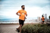 Foto do Thiago em movimento, correndo. Ele veste uma camisa laranja com o nome da equipe estampado.