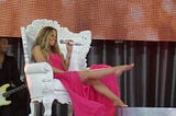 Mariah Carey, barefoot in a fancy pink dress holding a microphone and sitting in a big white throne