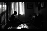 A black and white photograph shows a dimly lit room with a solitary figure sitting on the floor. The person is silhouetted against bright light coming through curtained windows, creating a stark contrast. suggesting a mood of sadness or contemplation. Shadows stretch across the wooden floor, and minimal furniture is visible, including what seems to be a small table. A few framed pictures hang on the walls. a strong sense of isolation, melancholy, and introspection through its dramatic lighting.