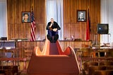 A stern robed judge at the top of an orange slide, holding a gavel, his feet facing us. He is in a courtroom. On the walls behind him hang portraits of Lina Khan and John Sherman. Image: Harrison Keely (modified) https://commons.wikimedia.org/wiki/File:The_Clay_County_Courthouse_courtroom_in_2007.jpg CC BY 4.0 https://creativecommons.org/licenses/by/4.0/deed.en — DocteurCosmos (modified) https://commons.wikimedia.org/wiki/File:Playground_slide_close-up.jpg CC BY 3.0 https://creativecommons