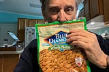 A man holding a bag of Blue Diamond Whole Natural Almonds in what appears to be a kitchen. The package has a green border and displays “Irresistible Snacking!” at the top. The almonds are visible through the packaging, and there’s an American Heart Association certification badge on the packet. The package also mentions “3g Net Carbs” on it. The person is holding the bag up near their face in what seems to be a home kitchen with wooden cabinets visible in the background.