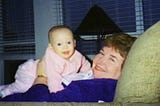 Beaming grandma, reclined in a comfy brown chair holding her baby granddaughter who is dressed in pink on her chest.