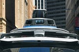 A self-driving car equipped with rooftop sensors and cameras navigates through an urban street canyon, flanked by high-rise buildings.